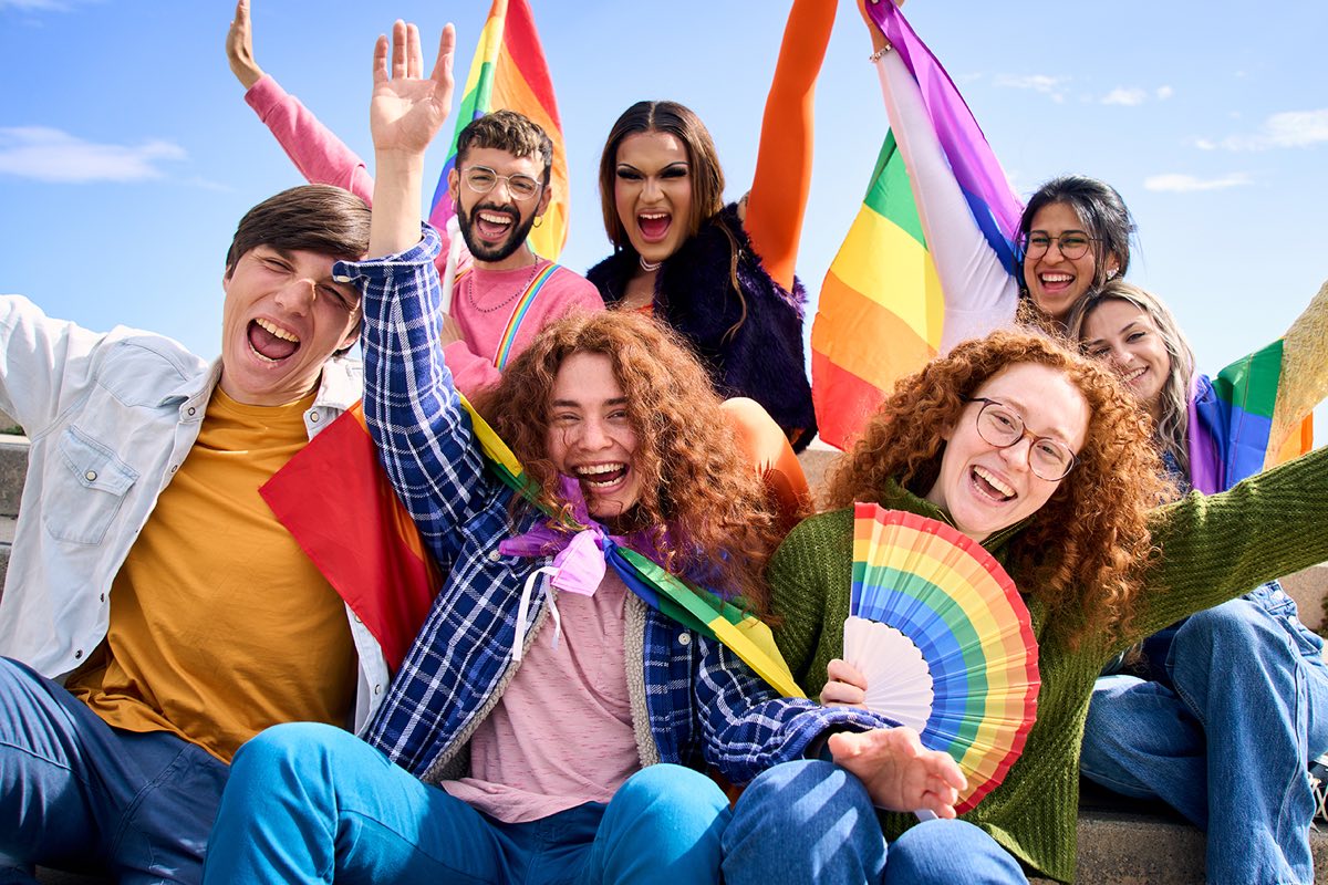 Seven friends laughing together on outdoor steps, holding Pride flags and a rainbow fan.