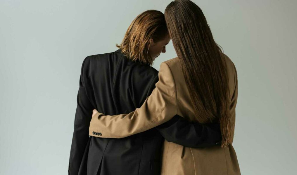 Back View Of Lesbian Women In Black And Beige Blazers Embracing Isolated On Grey.
