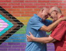 Mature gay couple embrace in front of a brick wall painted with