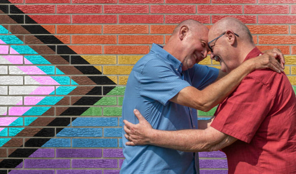 Mature gay couple embrace in front of a brick wall painted with