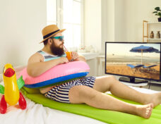 Funny man with inflatable beach toys sipping cocktail and watching travel TV show in lockdown