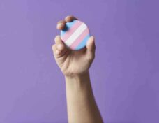 Hand Raising A Trans Flag Badge Or Pin On A Purple Background. Concepts Of Identity Pride, Gender Di