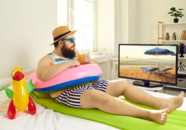 Funny man with inflatable beach toys sipping cocktail and watching travel TV show in lockdown