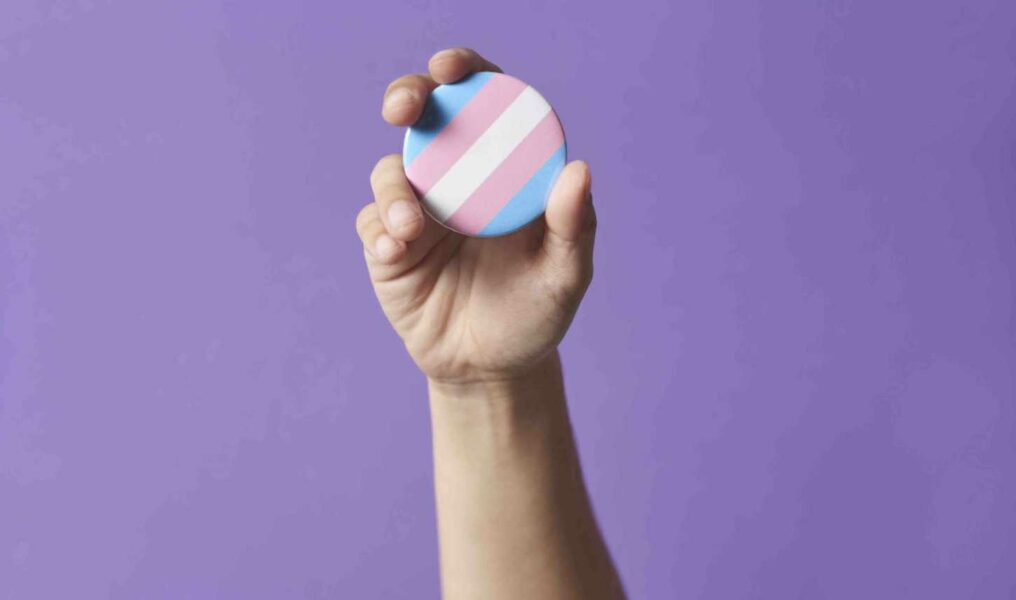 Hand Raising A Trans Flag Badge Or Pin On A Purple Background. Concepts Of Identity Pride, Gender Di