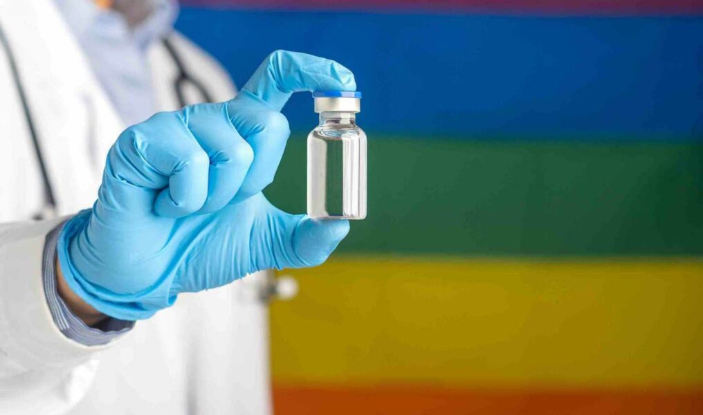 A Doctor Holding A Vaccine Bottle Against The Background Of The Rainbow Flag (lgbt). Vaccine For Imm
